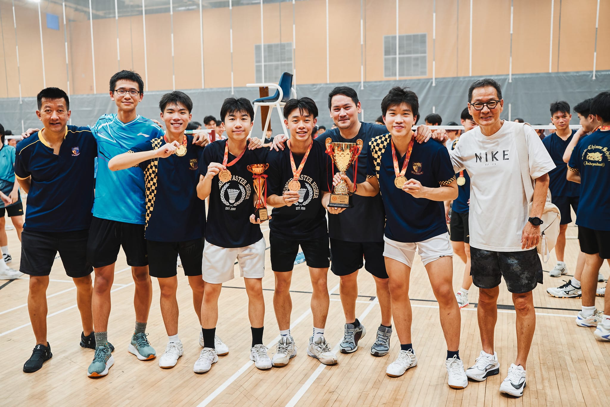 A badminton team in dark uniforms poses together with medals and trophies for a celebratory group photo.